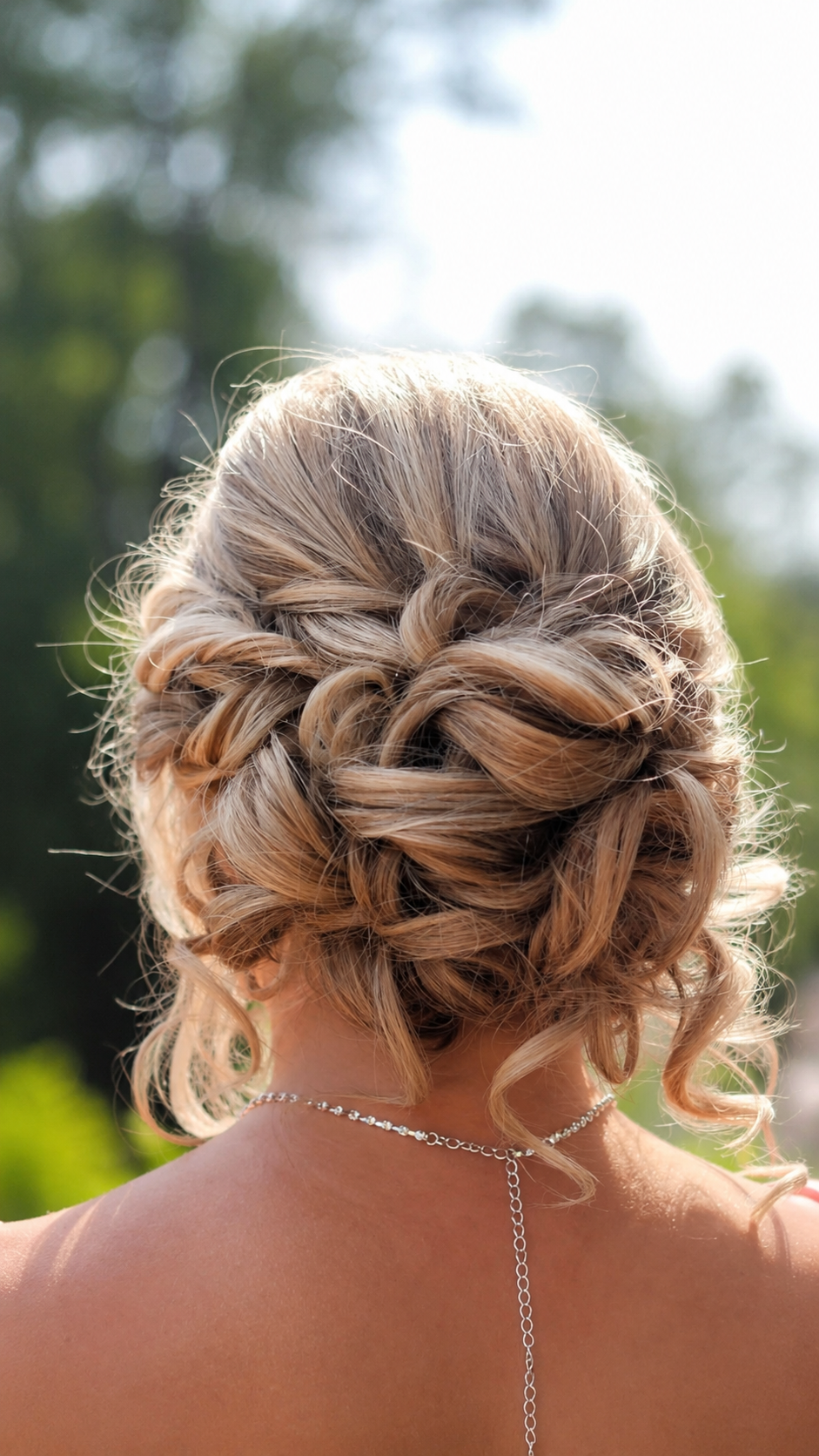 Back view of an elegant twisted updo with soft curls and braided detail, outdoor formal setting